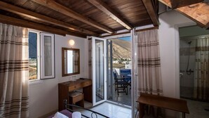 A room with a wooden ceiling, a bed, a desk, a chair, a table, a mirror, and a sliding glass door leading to a balcony with a view of a mountain and buildings. - Petra Nera (Santorini)
