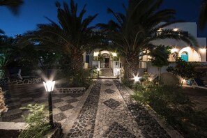 A nighttime view of a building with a stone pathway leading to the entrance, surrounded by palm trees and illuminated by street lamps. - Petra Nera (Santorini)
