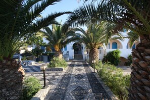 A cobblestone pathway leading to a building with a white facade and blue doors, surrounded by palm trees. - Petra Nera (Santorini)