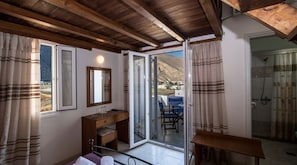 A room with a wooden ceiling, a bed, a desk, a chair, a table, a mirror, and a sliding glass door leading to a balcony with a view of a mountain and buildings. - Petra Nera (Santorini)