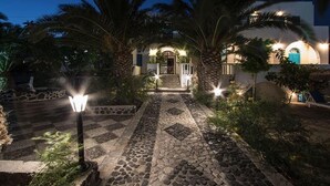A well-lit pathway leading to a building with a stone-tiled entrance, surrounded by palm trees and a stone wall. - Petra Nera (Santorini)