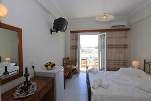 A hotel room with a bed, a desk with a fruit bowl, a TV, and a balcony with a table and chairs. - Petra Nera (Santorini)