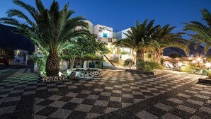 A hotel courtyard with palm trees, a checkered floor, and a building with a sign reading "Petra Mesa". - Petra Nera (Santorini)
