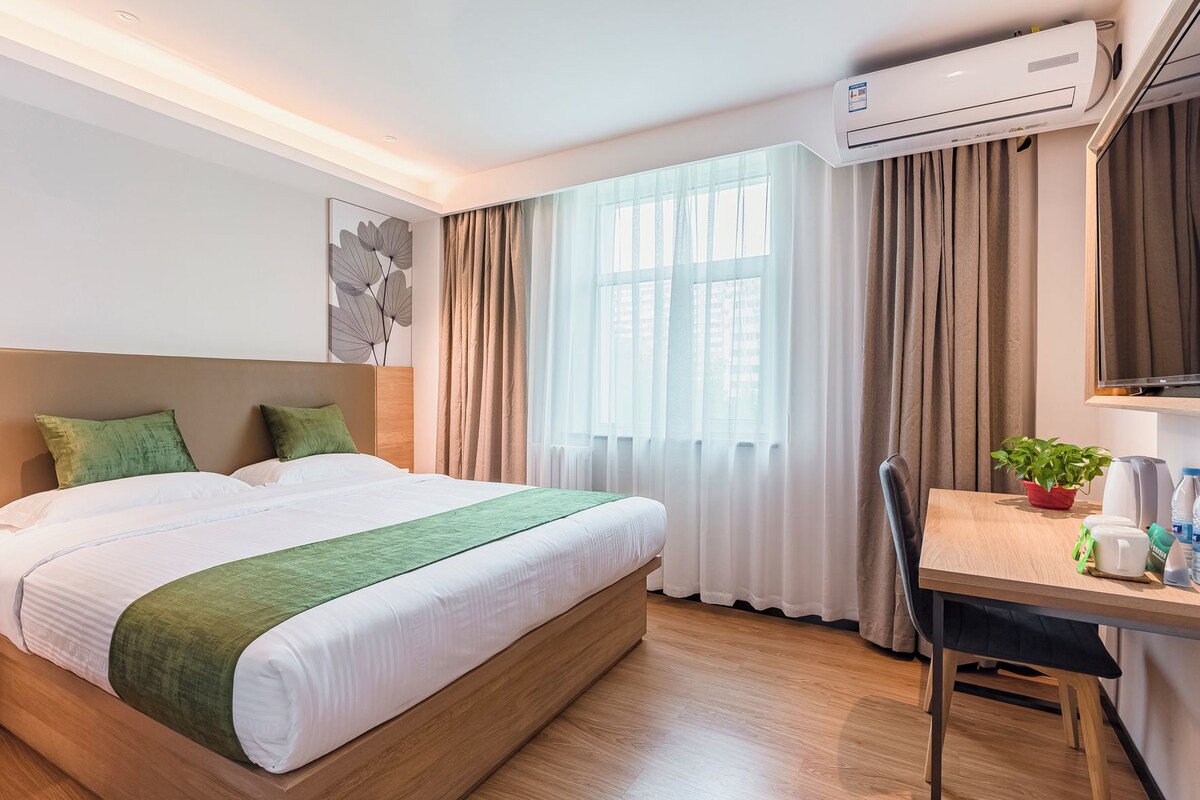 A hotel room with a bed, a desk, an air conditioning unit, a window with curtains, and a potted plant.