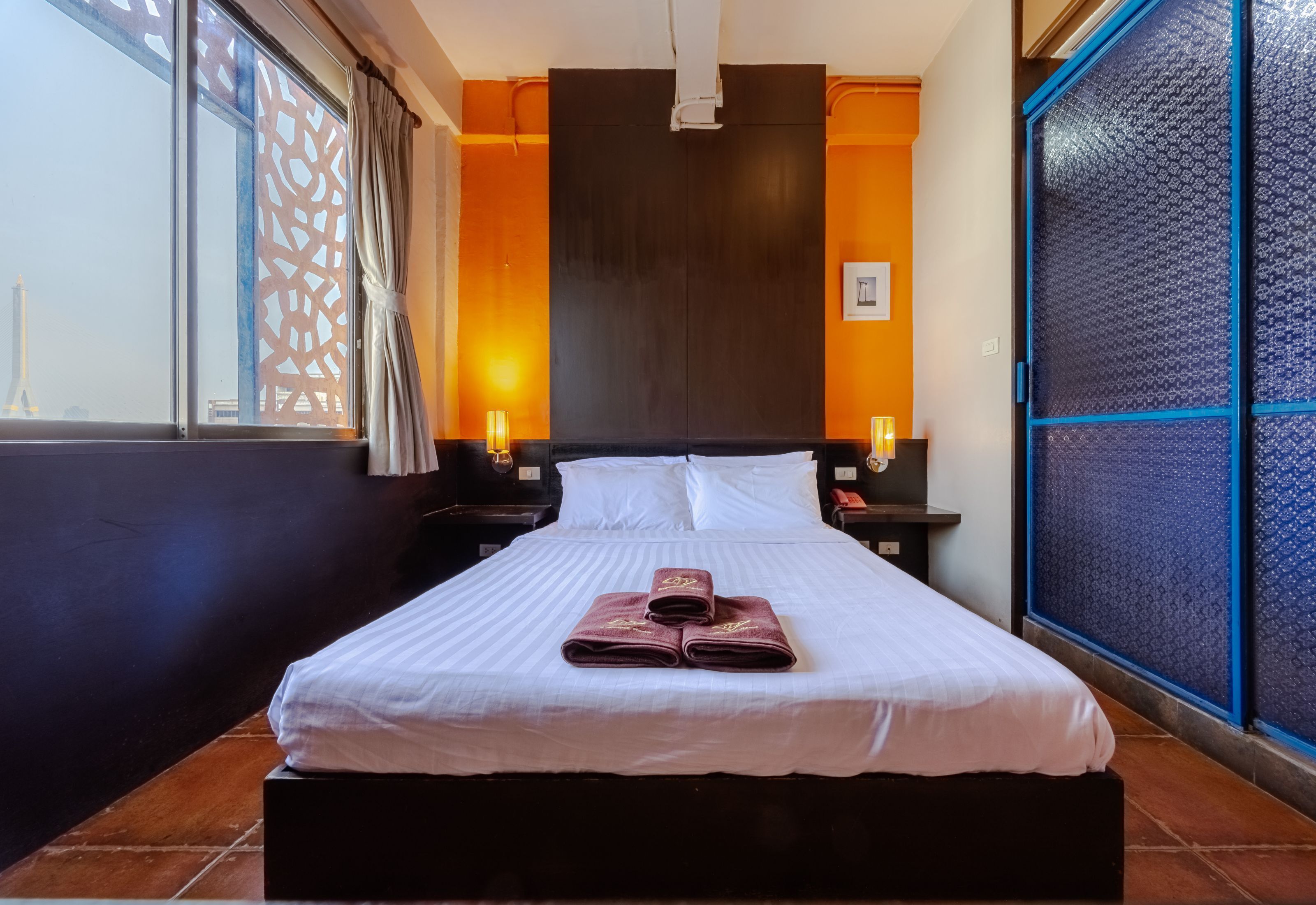 A neatly made bed with white linens and two folded towels, a dark wooden headboard, and a view of the Eiffel Tower through a window with a decorative screen.