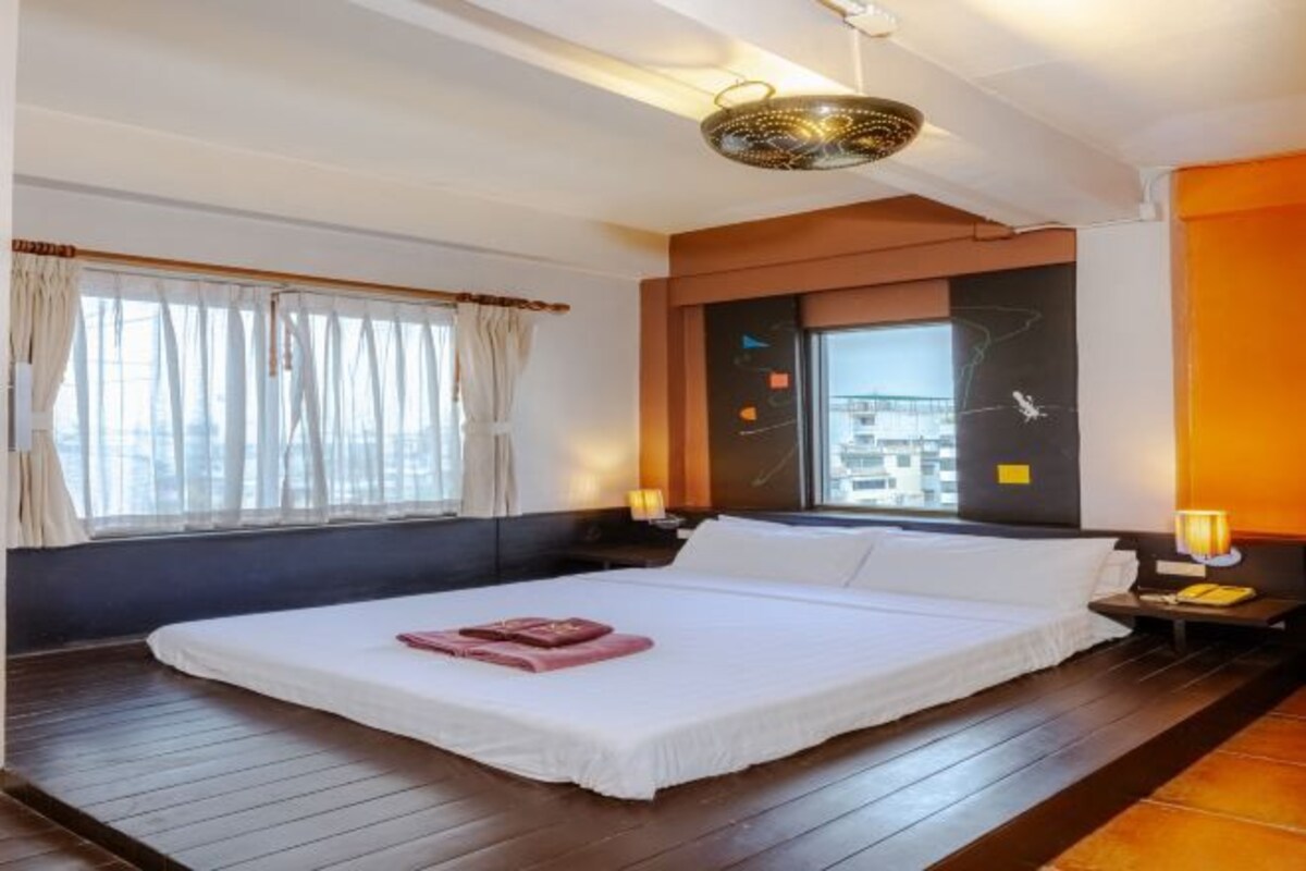 A hotel room with a large bed, a window with curtains, a bedside table with a lamp, and a modern ceiling light fixture.