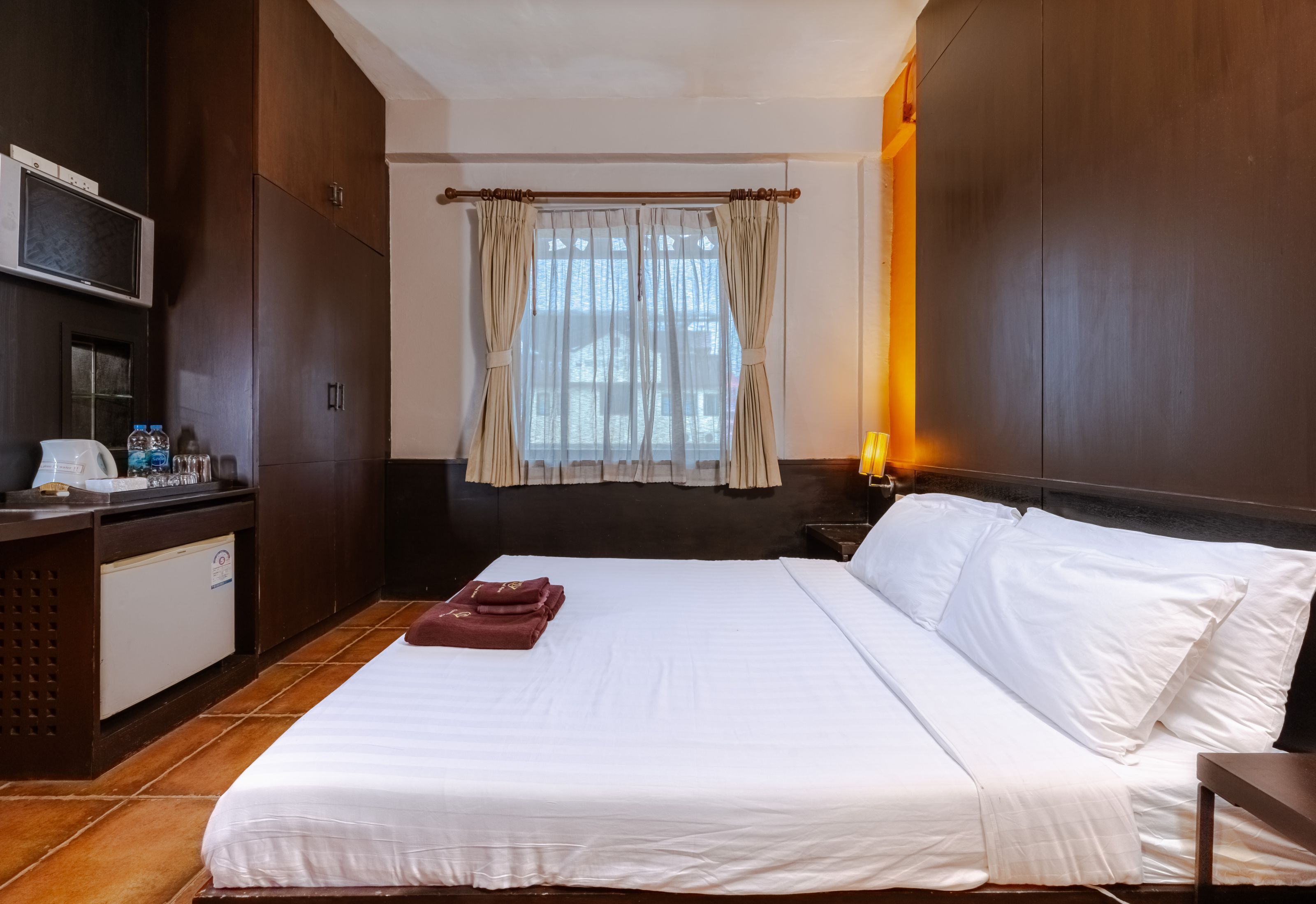 A hotel room with a bed, a desk with a microwave, a refrigerator, a window with curtains, and a nightstand with a lamp.