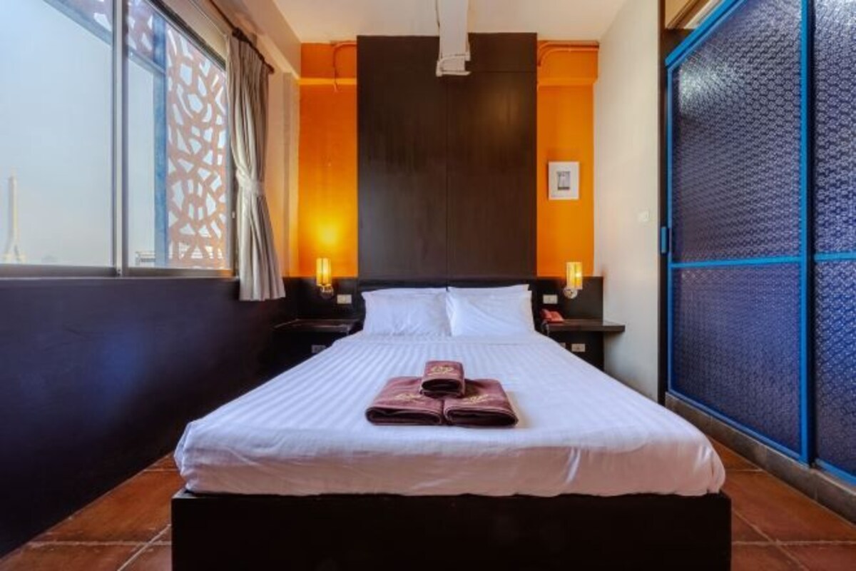A hotel room with a bed, two bedside tables, a window with a view, and a door with a textured glass panel.