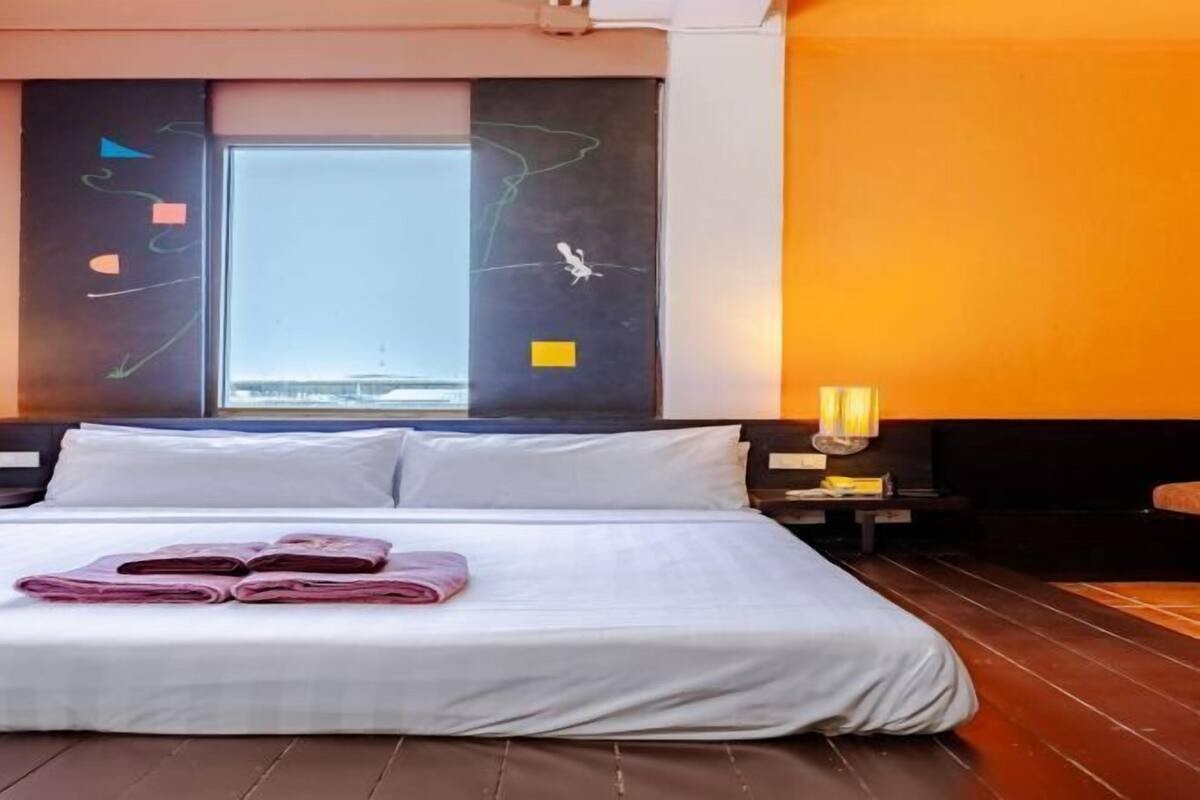 A hotel room with a large bed, a window with a view, a bedside table with a lamp, and a wall with abstract art.