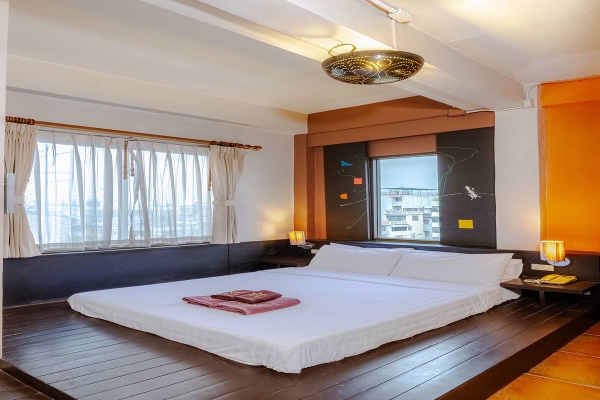 A hotel room with a large bed, wooden flooring, a window with curtains, a wall-mounted light, and a view of the city.