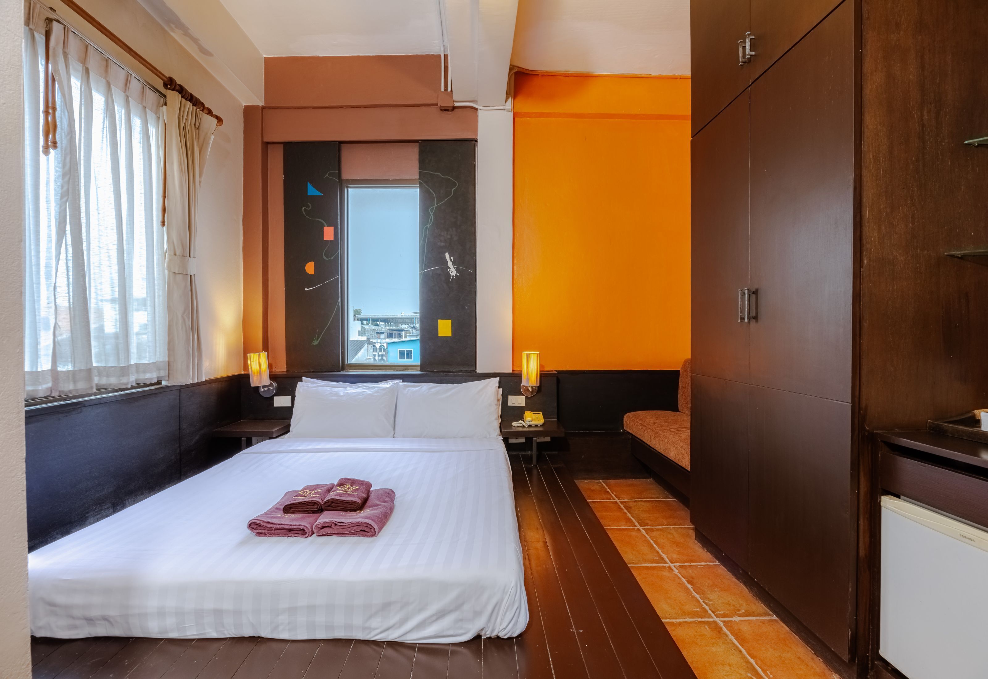 A hotel room with a large bed, a nightstand, a wardrobe, and a view of the city through a window.