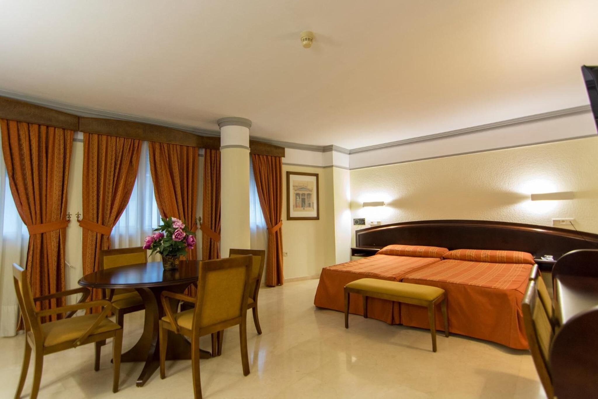 A hotel room with a dining table, chairs, a bench, a bed with an orange bedspread, and a framed picture on the wall.