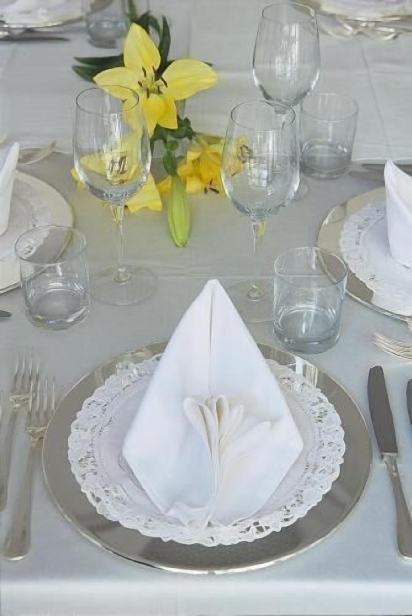 Una mesa dispuesta con servilletas blancas dobladas en forma de cono, copas transparentes y un arreglo floral amarillo.
