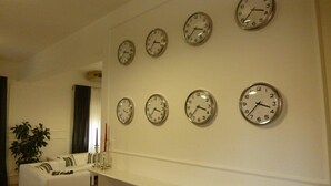 A room with a white wall and multiple clocks. There is a white sofa with a striped pillow and a plant on the left side. - Hotel Amici (Agrigento)