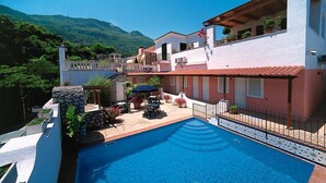 A pool area with a clear view of a mountain in the background, a patio with outdoor seating, and a building with a red-tiled roof. - Hotel Il Nespolo (Casamicciola Terme)