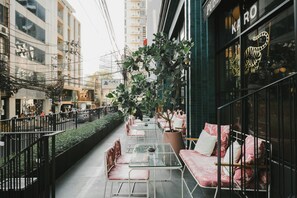 Un café al aire libre con mesas y sillas de vidrio, rodeado por una cerca de metal y plantas en macetas.