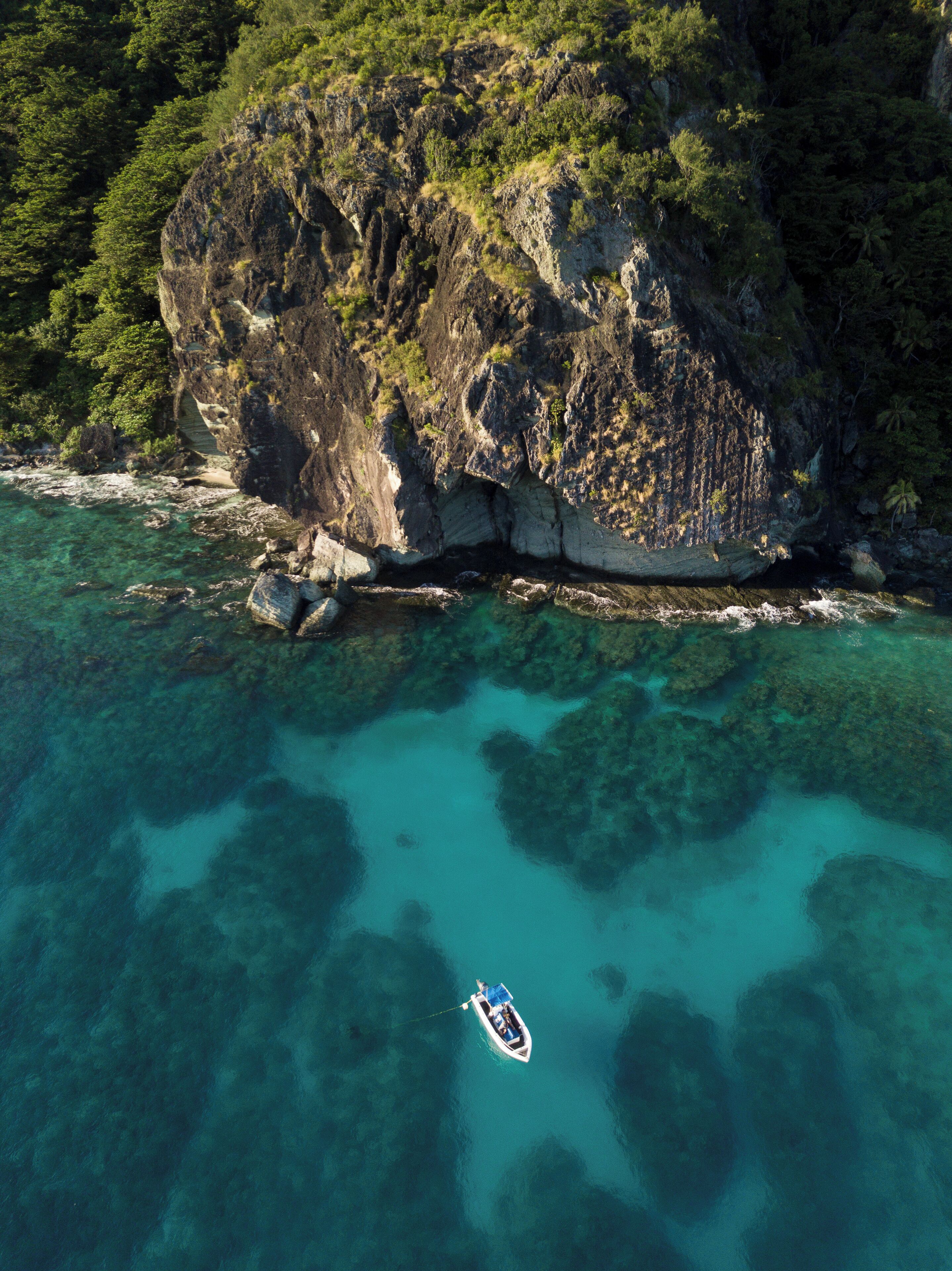 An aerial view of a coastal area with clear turquoise waters, a rocky cliff, and a small boat anchored near the shore.