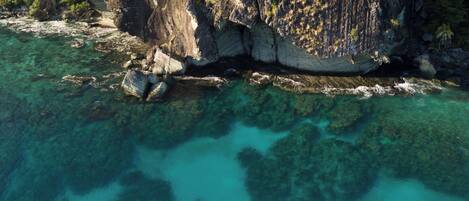 An aerial view of a coastal area with clear turquoise waters, a rocky cliff, and a small boat anchored near the shore.