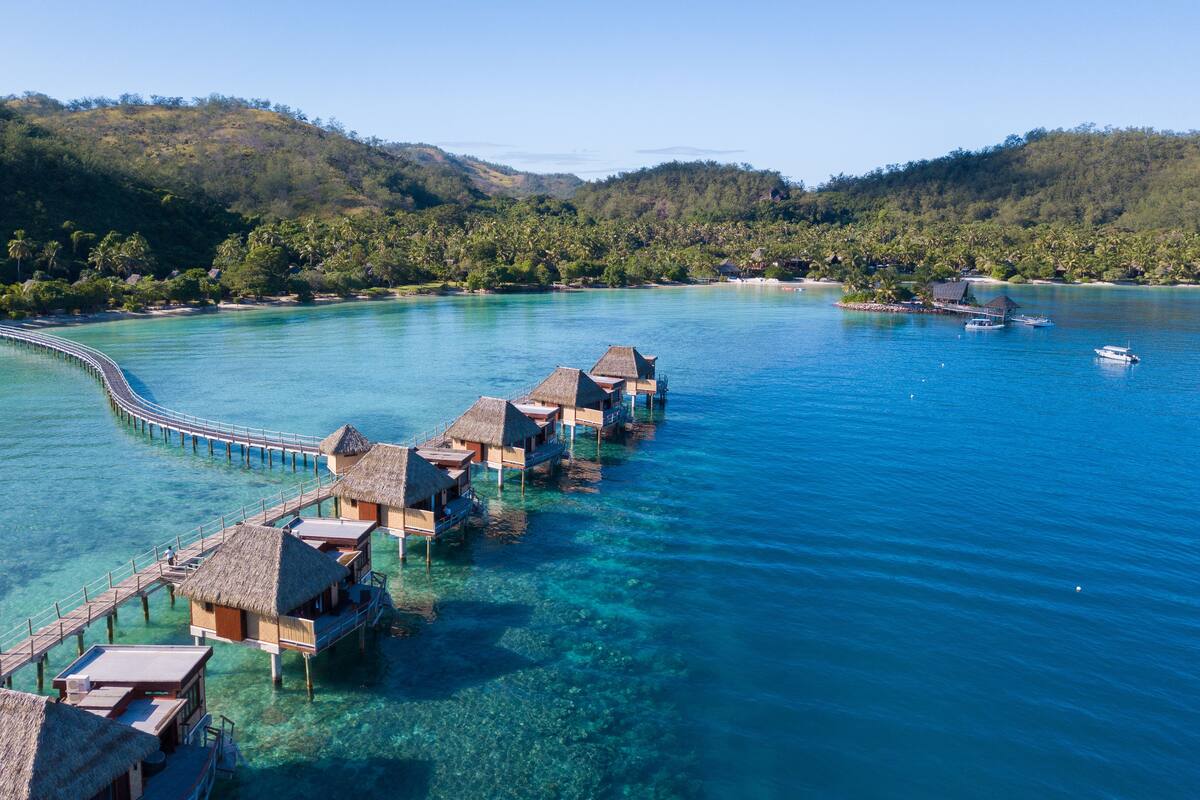 Cabañas sobre el agua conectadas por una pasarela de madera, rodeadas de aguas turquesas cristalinas y exuberante vegetación.