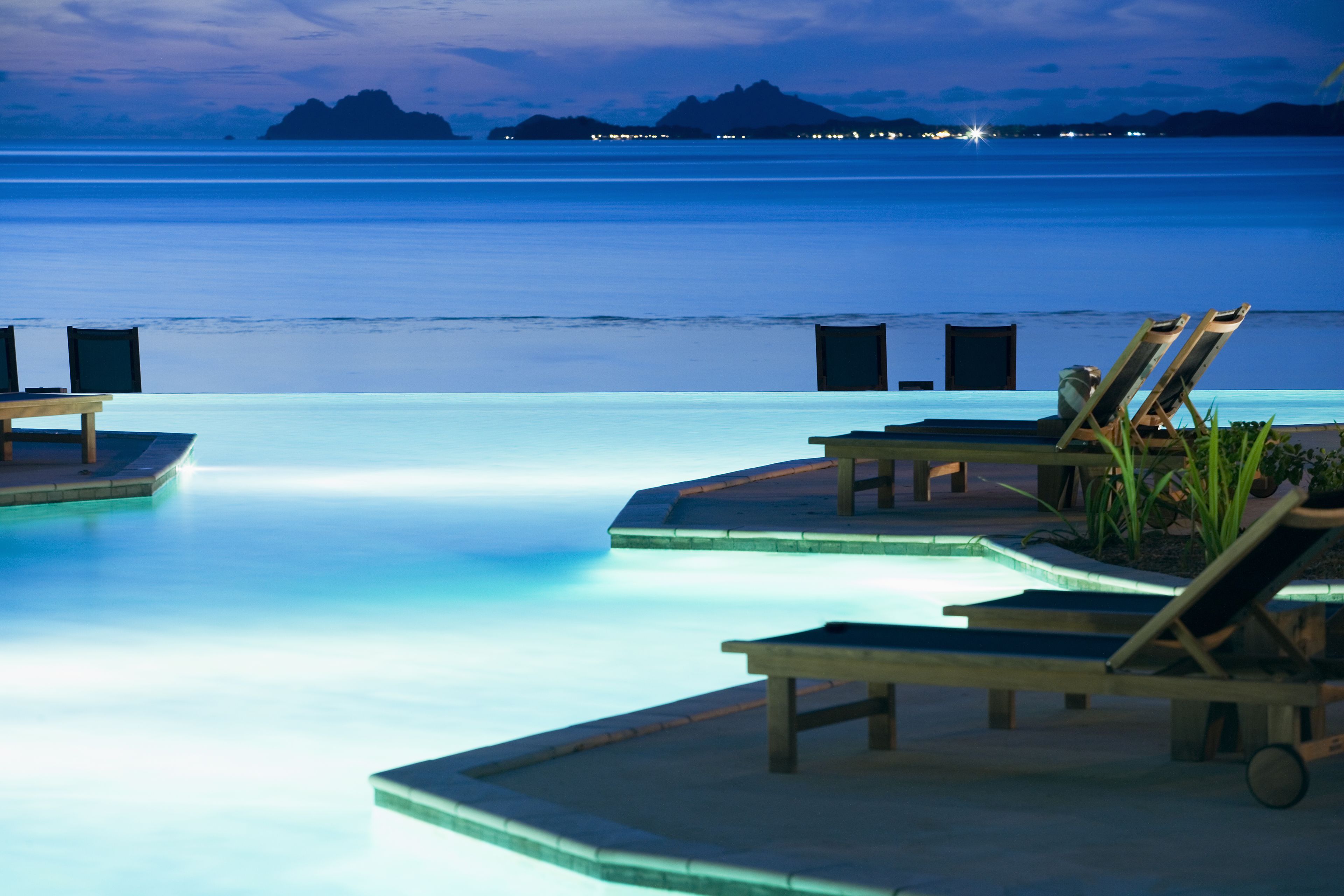 Zona de piscina con sillones de descanso y vista al océano al atardecer.