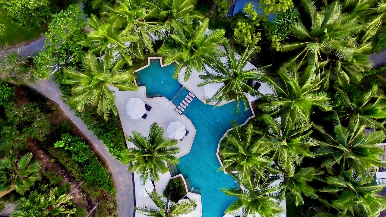 Eine Vogelperspektive zeigt ein Resort mit einem zentralen Schwimmbecken, umgeben von Palmen und Liegestühlen.