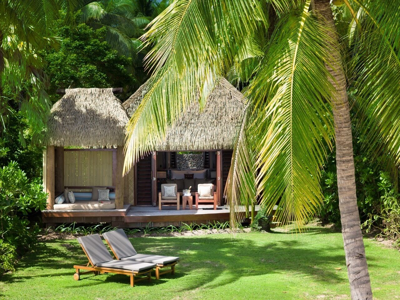 A thatched-roof structure with a seating area and a small table, surrounded by lush greenery and palm trees.