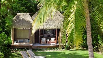 A thatched-roof structure with a seating area and a small table, surrounded by lush greenery and palm trees.