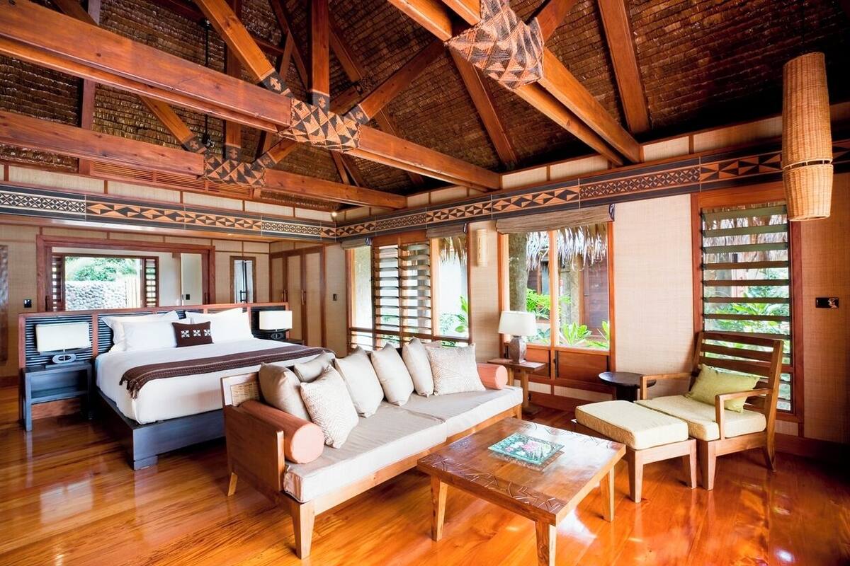 Habitación amplia con una cama grande, un sofá y una silla. Vigas de madera en el techo y una ventana con vistas a la vegetación.