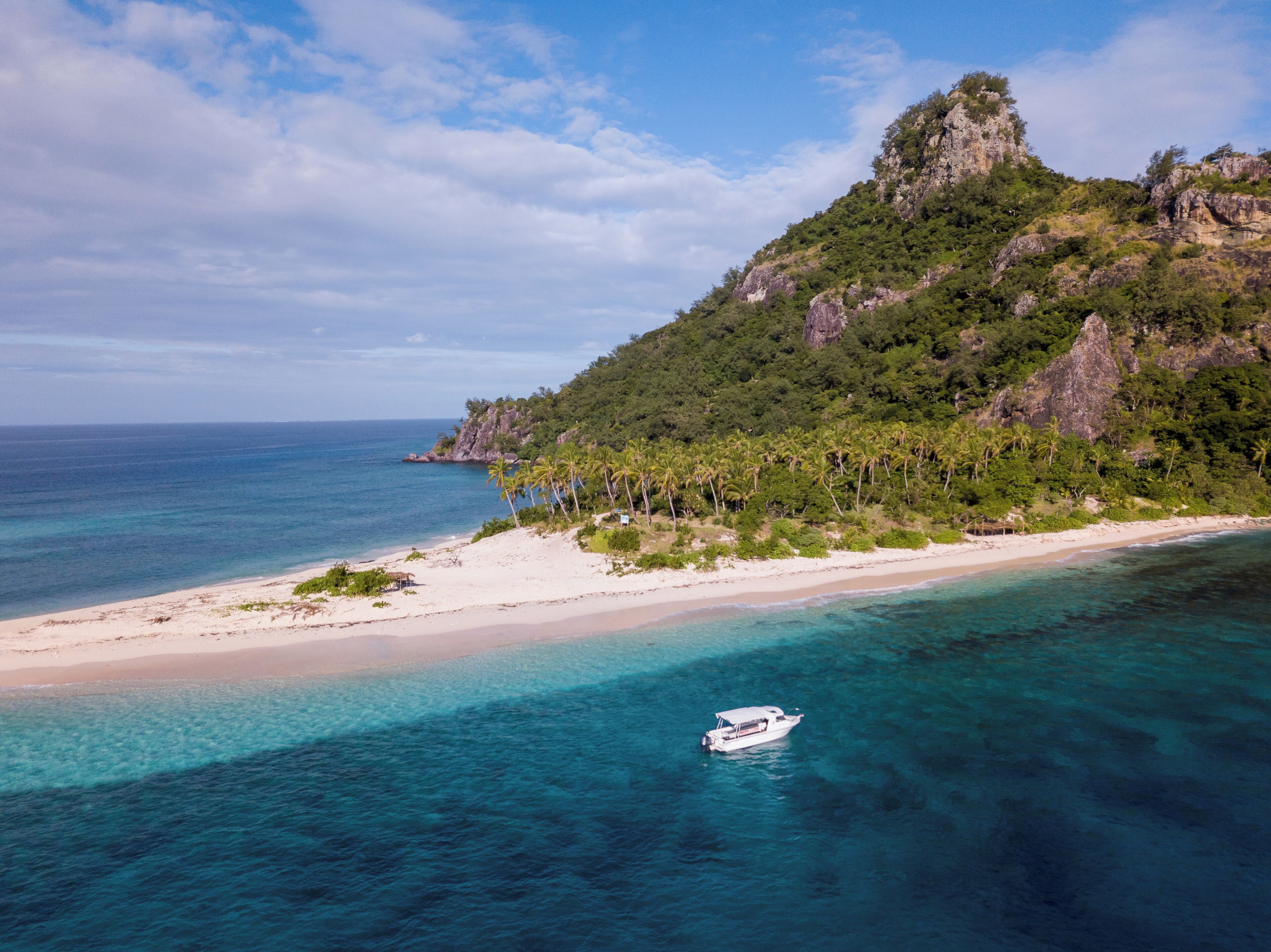 A beach with clear turquoise waters, a boat, and a mountainous landscape with dense vegetation.