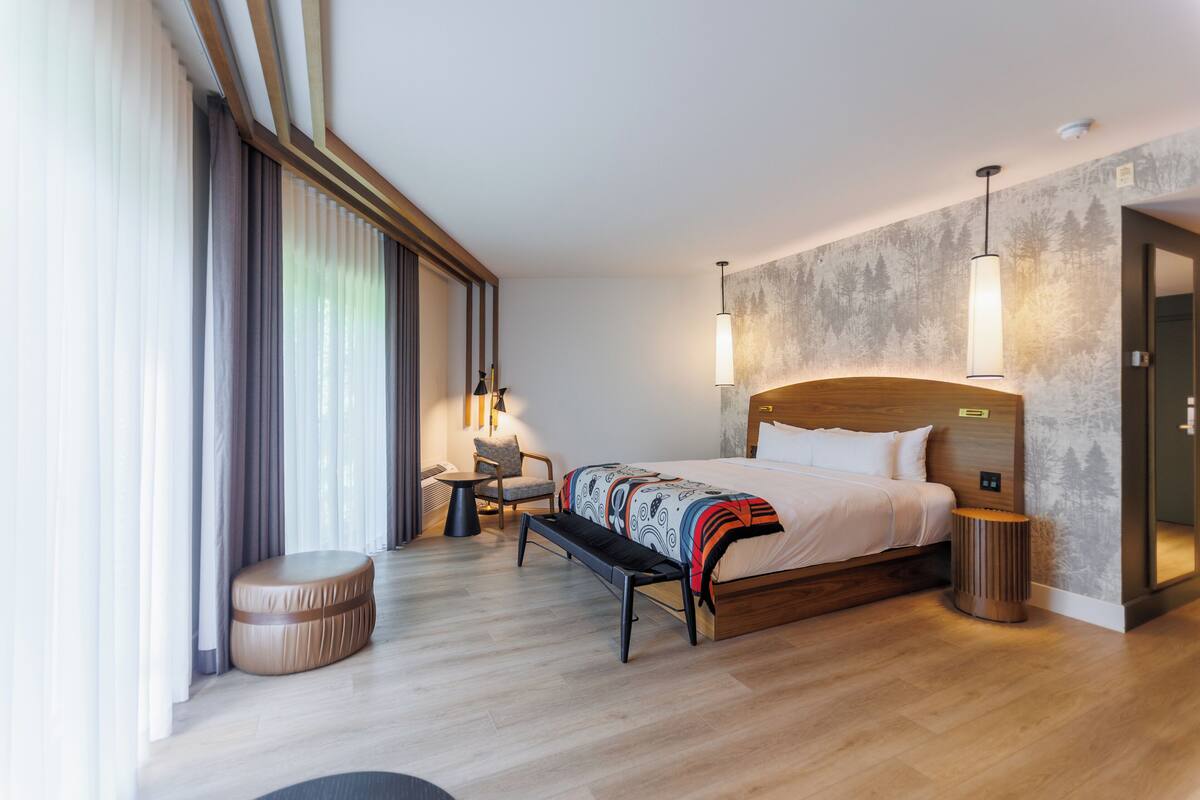 Habitación de hotel moderna con una cama grande, un banco, una mesita pequeña y una silla. Cuenta con un ventanal con cortinas, una lámpara de pared y un taburete de madera redondo.