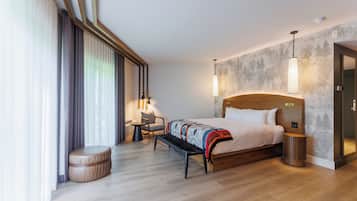 Habitación de hotel moderna con una cama grande, un banco, una mesita pequeña y una silla. Cuenta con un ventanal con cortinas, una lámpara de pared y un taburete de madera redondo.