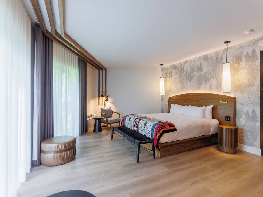Habitación de hotel moderna con una cama grande, un banco, una mesita pequeña y una silla. Cuenta con un ventanal con cortinas, una lámpara de pared y un taburete de madera redondo.