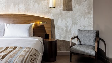 Una cama bien hecha con sábanas blancas, un cabecero de madera y una mesita de noche con una lámpara. Un sillón con estampado se encuentra al lado de la cama.