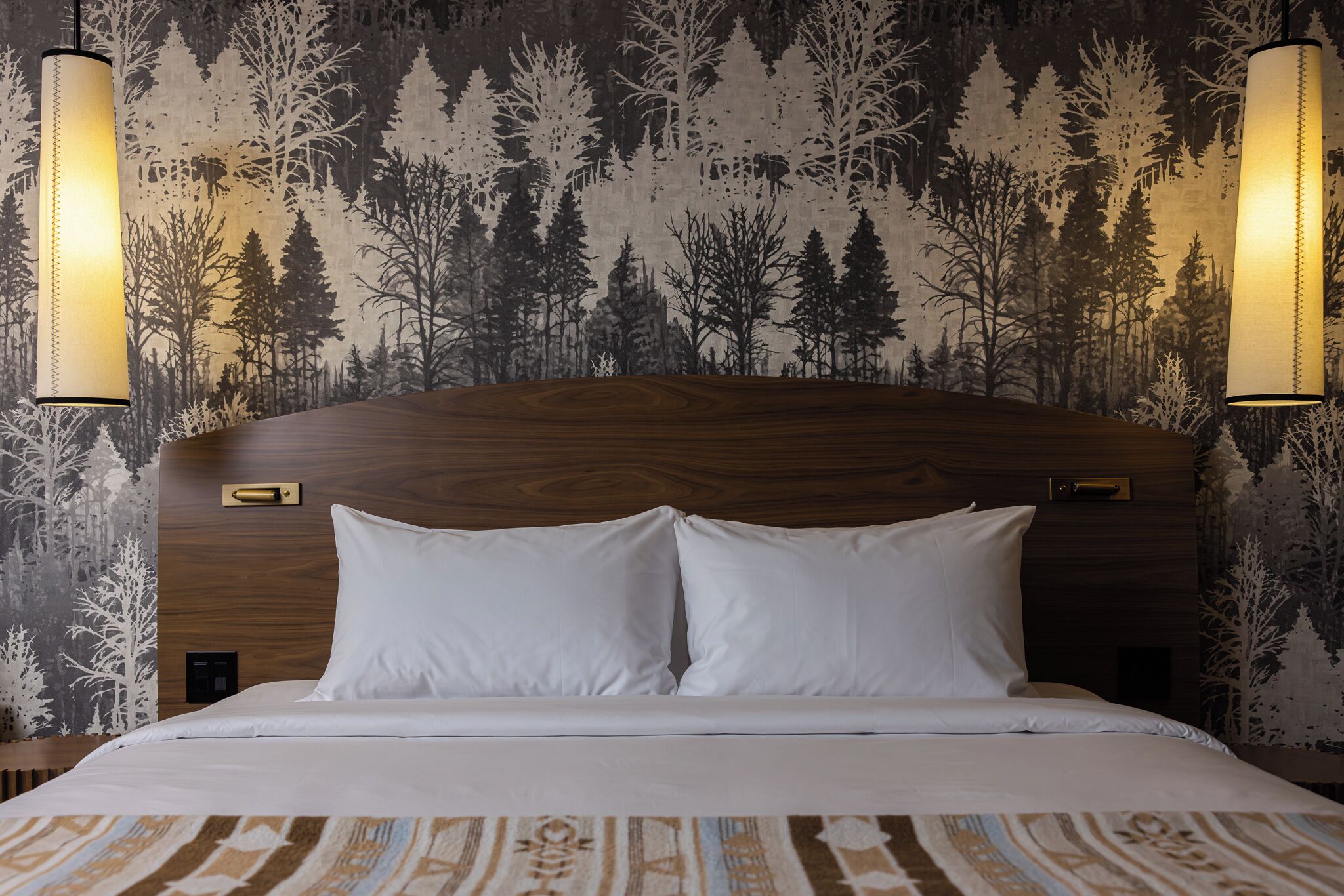 Ein Hotelzimmer mit einem Holzkopfende, zwei weißen Kissen, einer gemusterten Bettdecke und einem Wandpapier mit Waldbildern.
