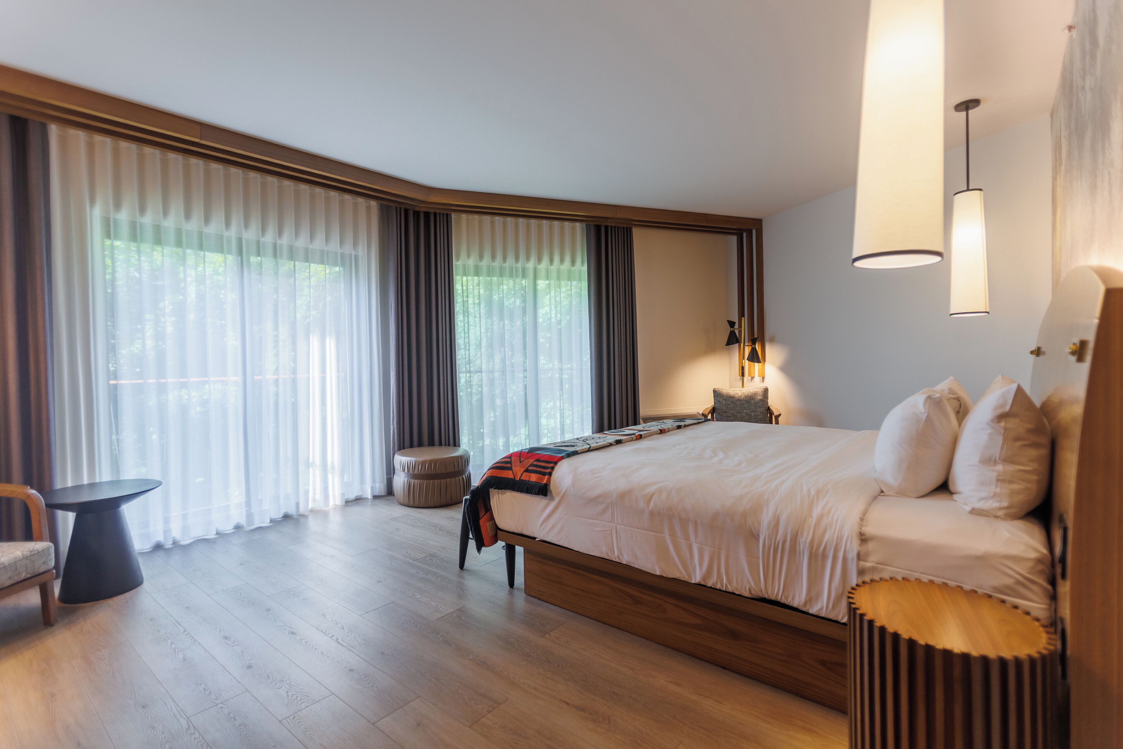 A modern hotel room with a large bed, a small round table, and a view of the outdoors through large windows.