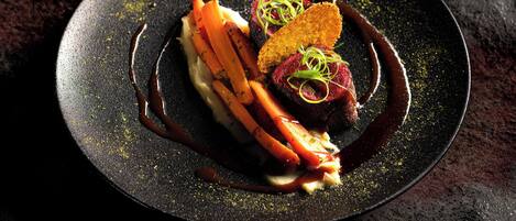 Un plato oscuro con un postre que incluye una porción de pastel, un trozo de carne y verduras, adornado con salsa.