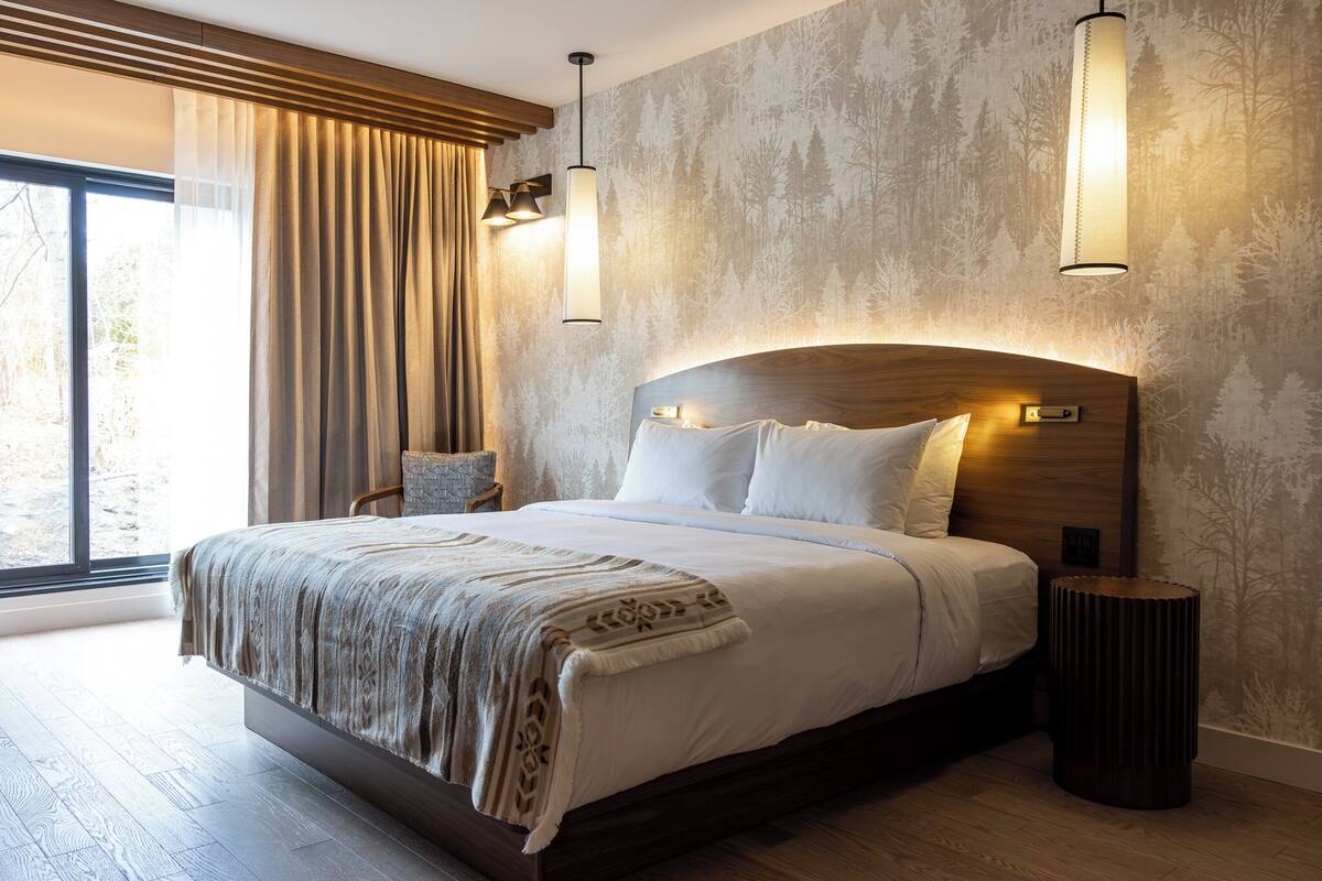 Una cama grande con cabecera de madera, dos lámparas de noche, una mesita auxiliar y vistas al exterior a través de un ventanal con cortinas.
