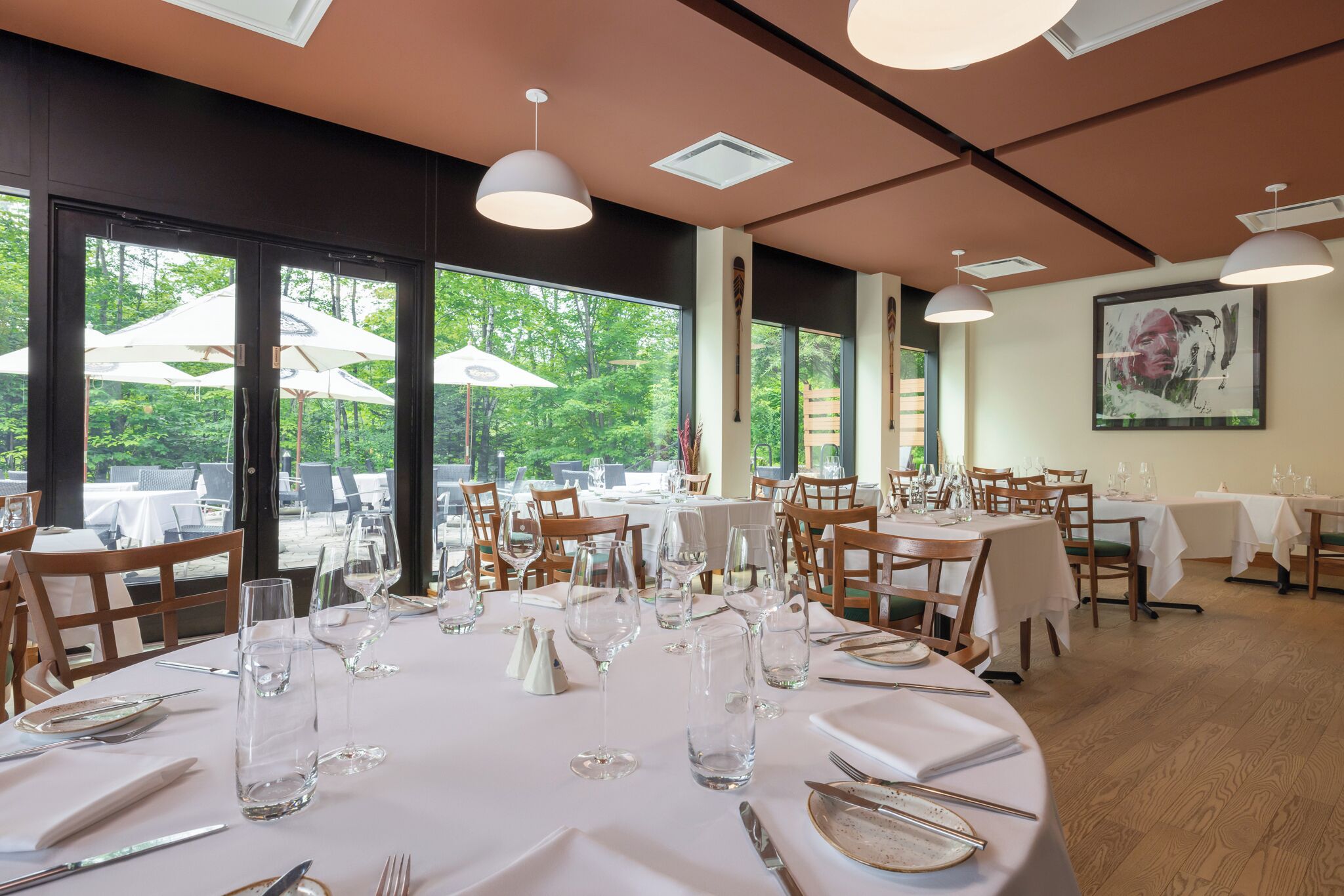 Un restaurant avec des tables rondes disposées pour le service, de grandes fenêtres donnant sur une terrasse ombragée par des parasols, et une vue sur la verdure.