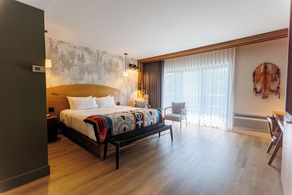 Una habitación de hotel moderna con una cama grande, un banco, una silla, un ventanal con cortinas y un calentador fijado a la pared.
