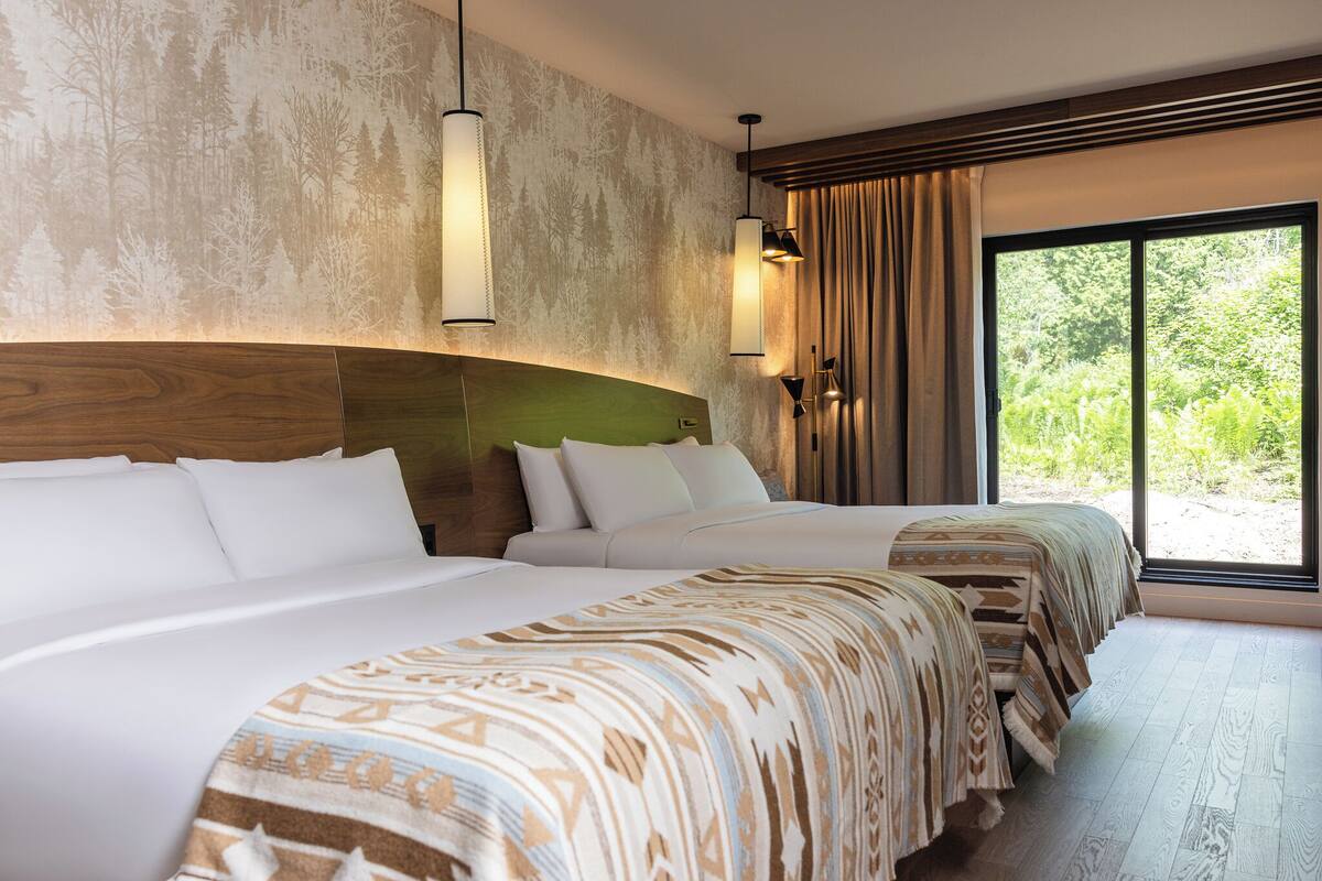 Habitación de hotel con dos camas, un ventanal grande y un mural con un paisaje de bosque en la pared.