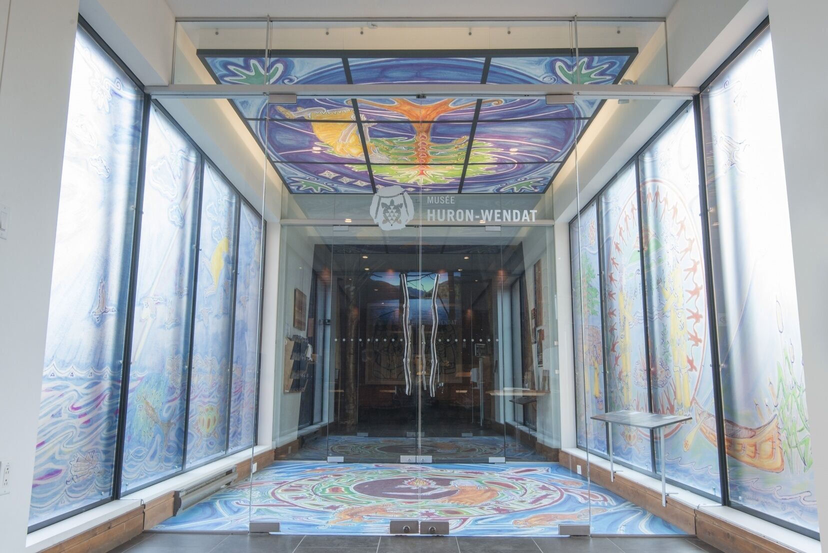 La entrada del museo Huron-Wendat con puertas de vidrio, un mural de techo colorido y un piso estampado.