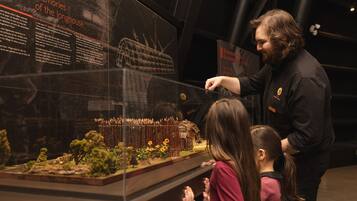 A museum exhibit with a detailed model of a longhouse, including a roof structure and surrounding landscape.