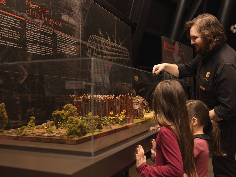 A museum exhibit with a detailed model of a longhouse, including a roof structure and surrounding landscape.