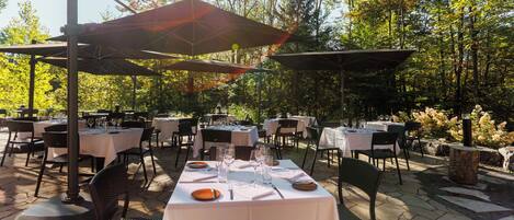 An outdoor dining area with tables set for two, surrounded by chairs and covered by umbrellas.