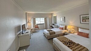 Quarto de hotel com duas camas, uma escrivaninha, uma cadeira, um abajur e uma janela com vista para edifícios.