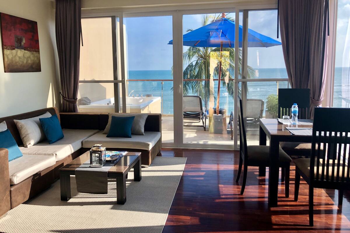 A modern living room with a sofa, coffee table, and chairs, a balcony with a view of the ocean, and a dining area with a table and chairs.
