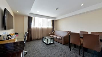 A hotel room with a flat-screen TV, a sofa, a coffee table, and a desk with a computer and a tray of items.