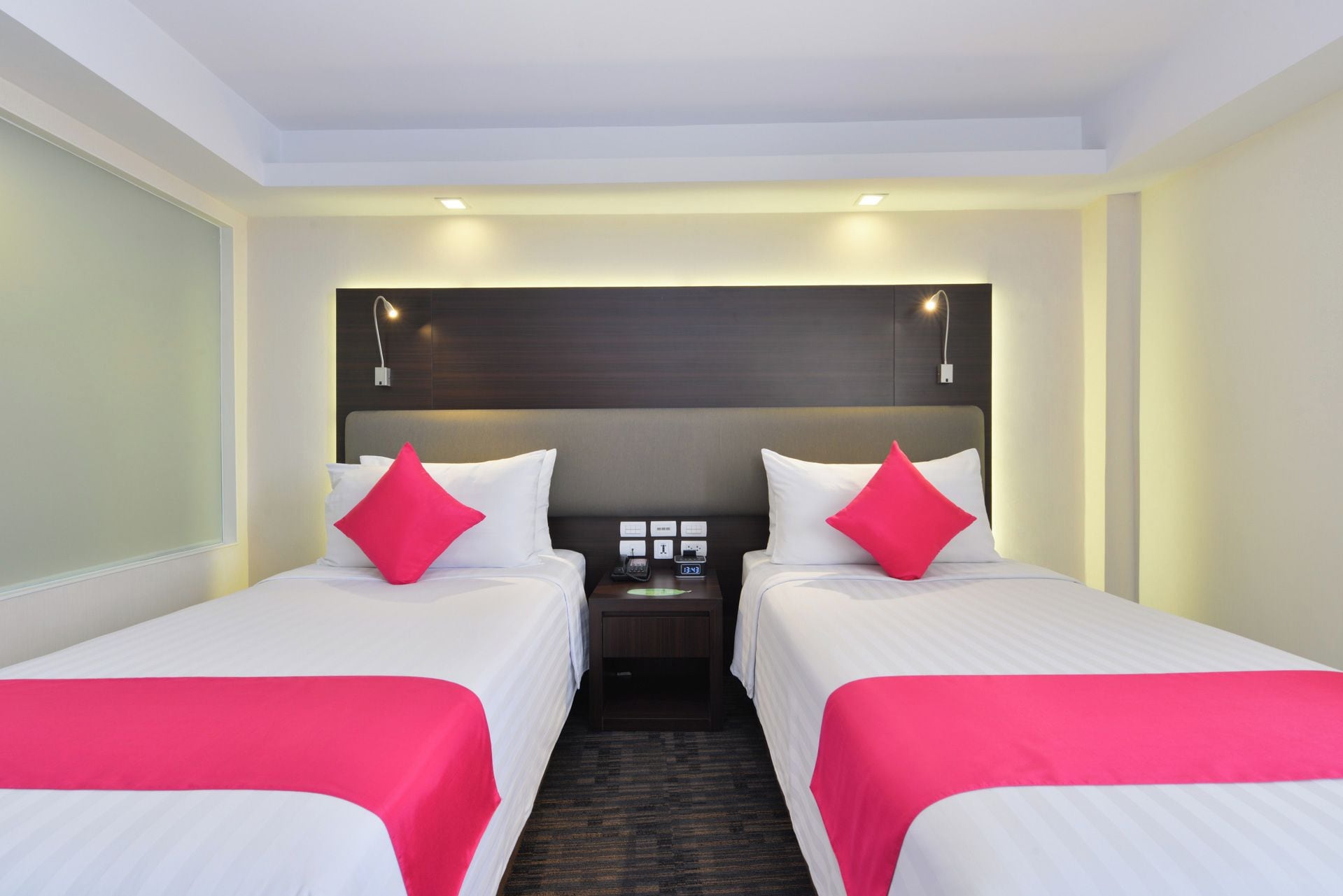 Een hotelkamer met twee bedden, elk met wit beddengoed en rode kussens, een nachtkastje met een telefoon en een klok, en een donker hoofdeinde.
