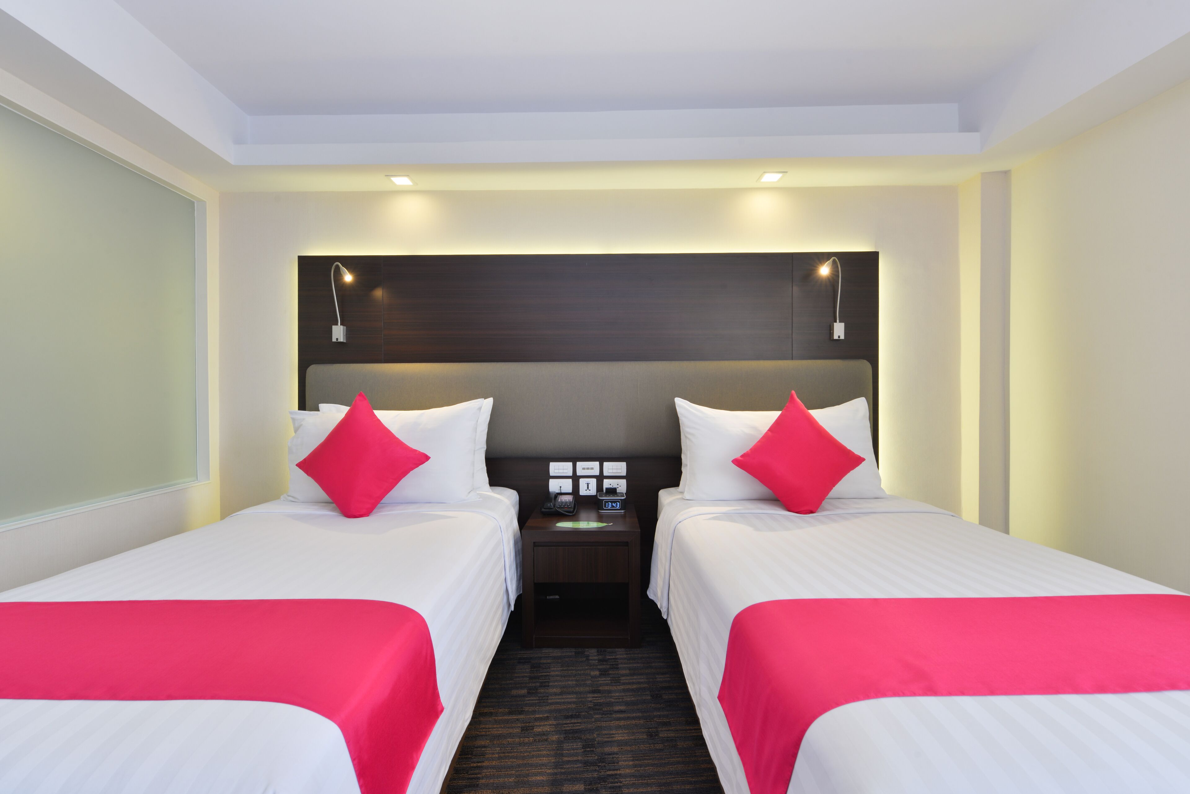 A hotel room with two single beds, each with a red accent pillow, a nightstand with a phone and a clock, and a dark headboard.
