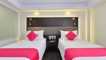 A hotel room with two single beds, each with a red accent pillow, a nightstand with a phone and a clock, and a dark headboard.